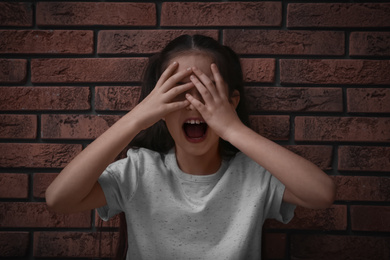 Scared little girl closing eyes with hands near brick wall. Child in danger
