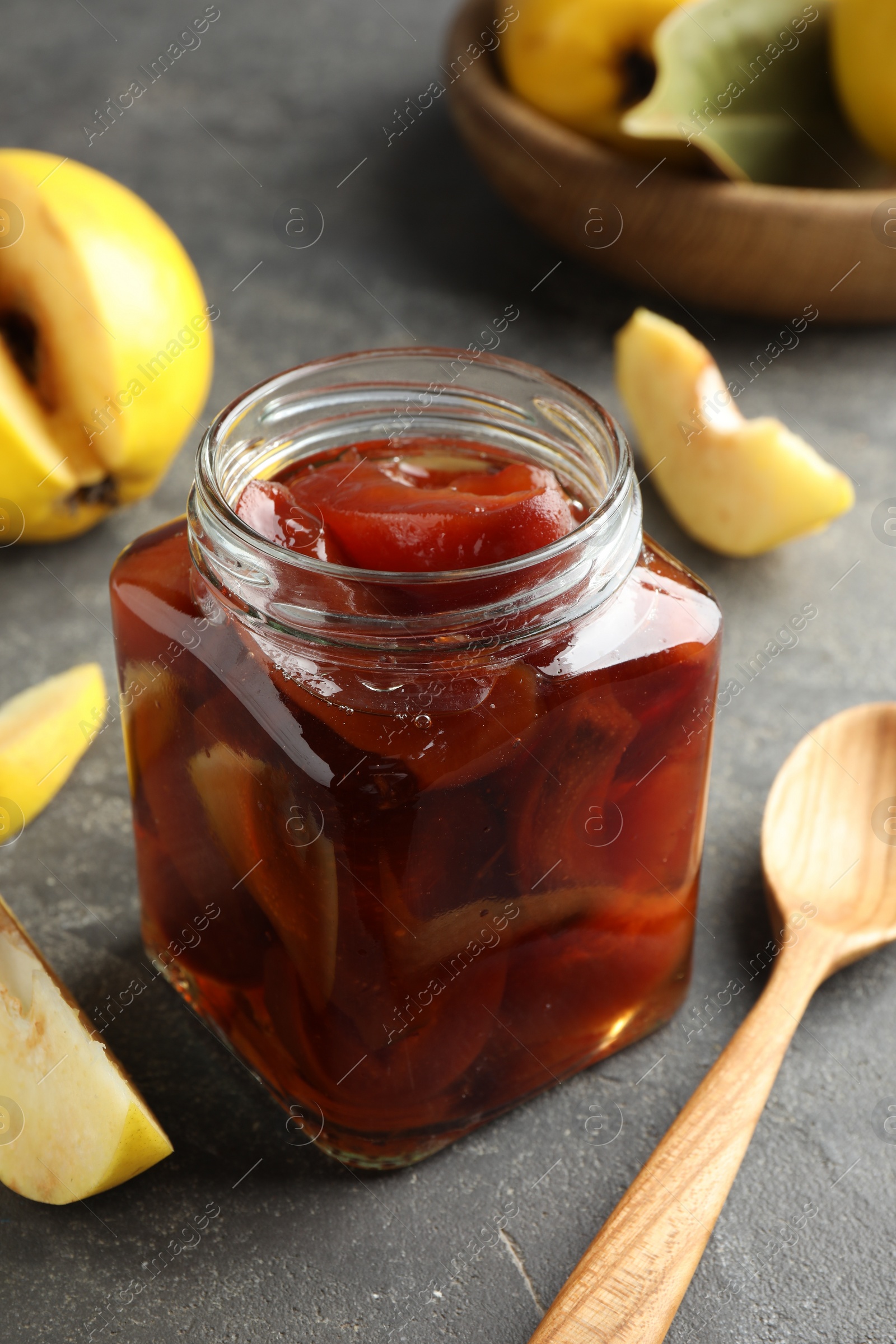 Photo of Tasty homemade quince jam in jar, spoon and fruits on grey textured table