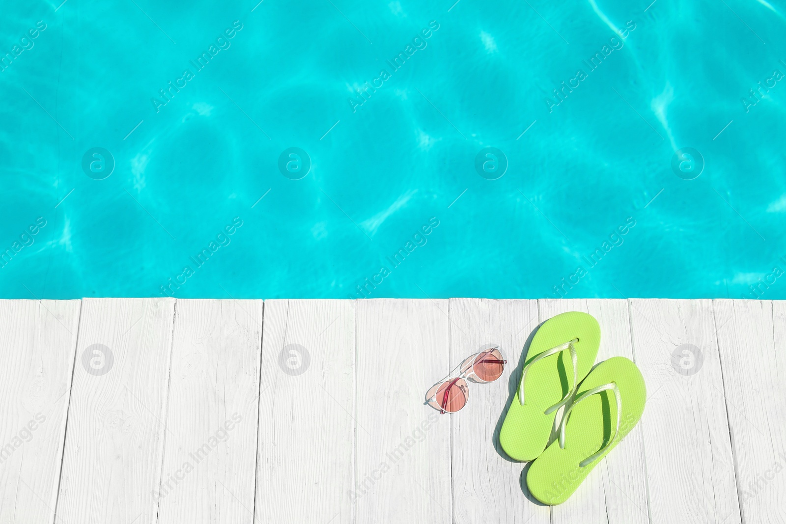 Photo of Slippers and sunglasses on wooden deck near swimming pool, top view with space for text. Beach accessory