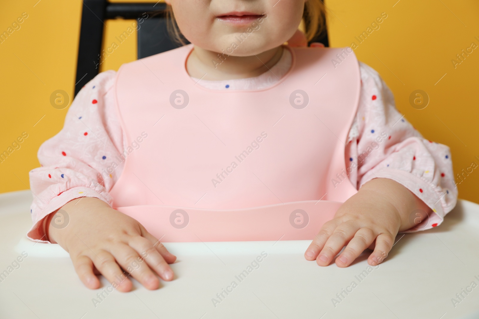 Photo of Cute little baby wearing bib in highchair on yellow background, closeup