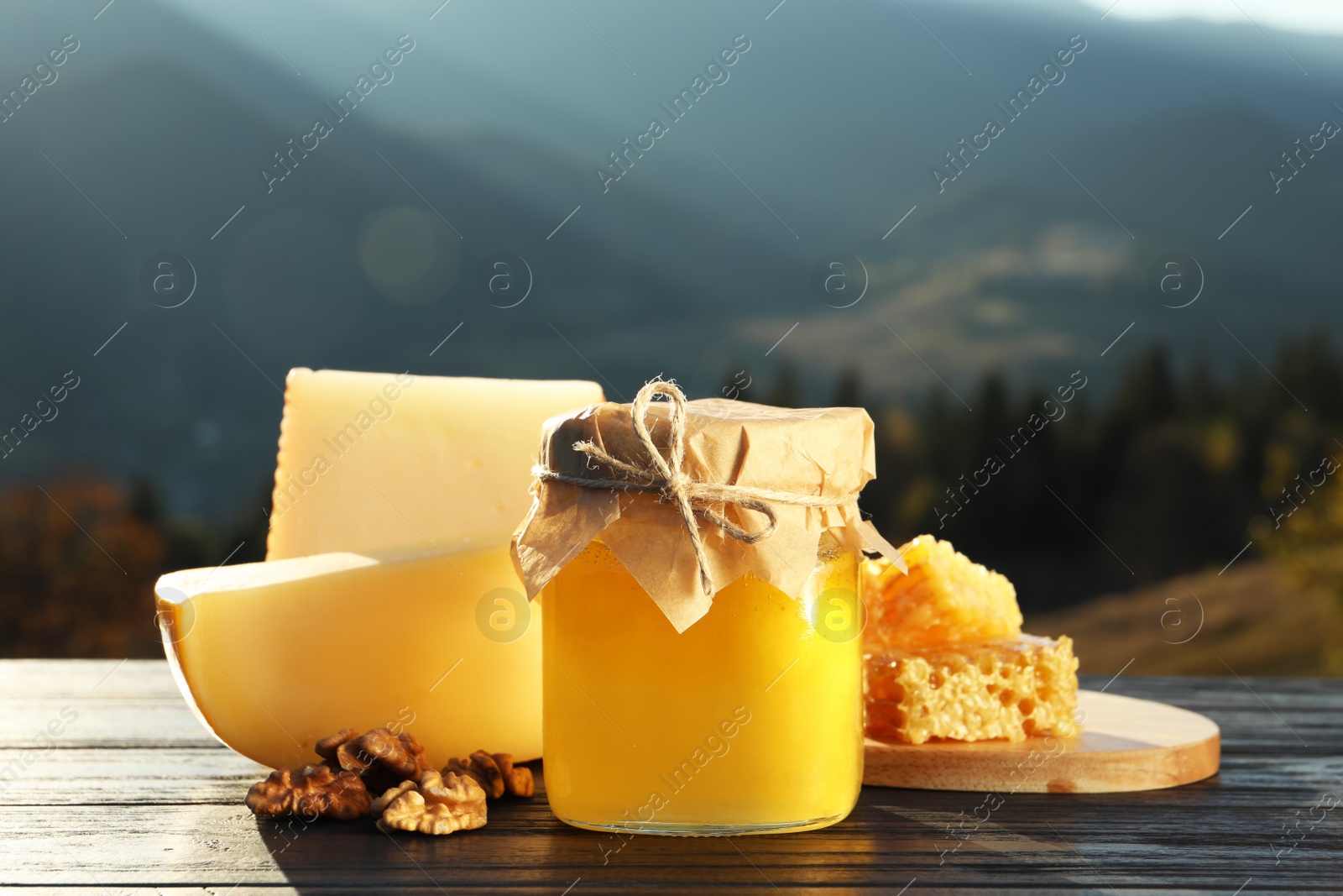 Photo of Fresh aromatic honey, combs, cheese and nuts on wooden table against mountain landscape