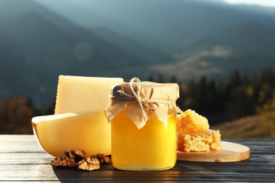 Photo of Fresh aromatic honey, combs, cheese and nuts on wooden table against mountain landscape