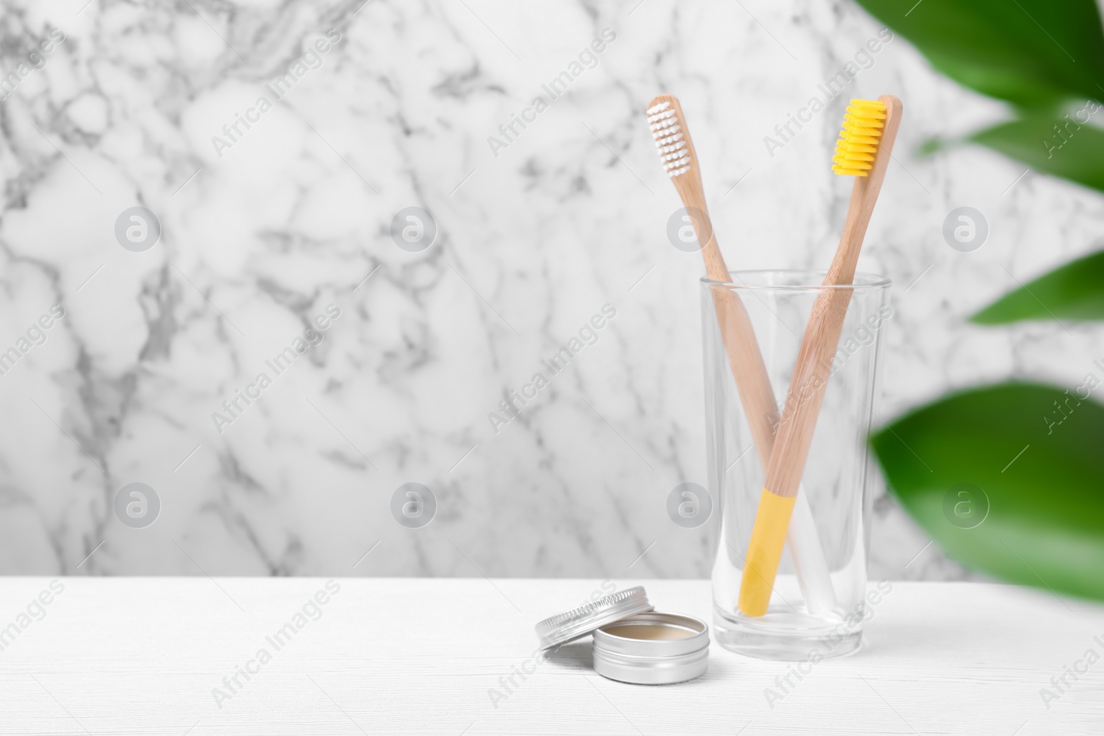 Photo of Glass with bamboo toothbrushes and paste on table against blurred background. Space for text