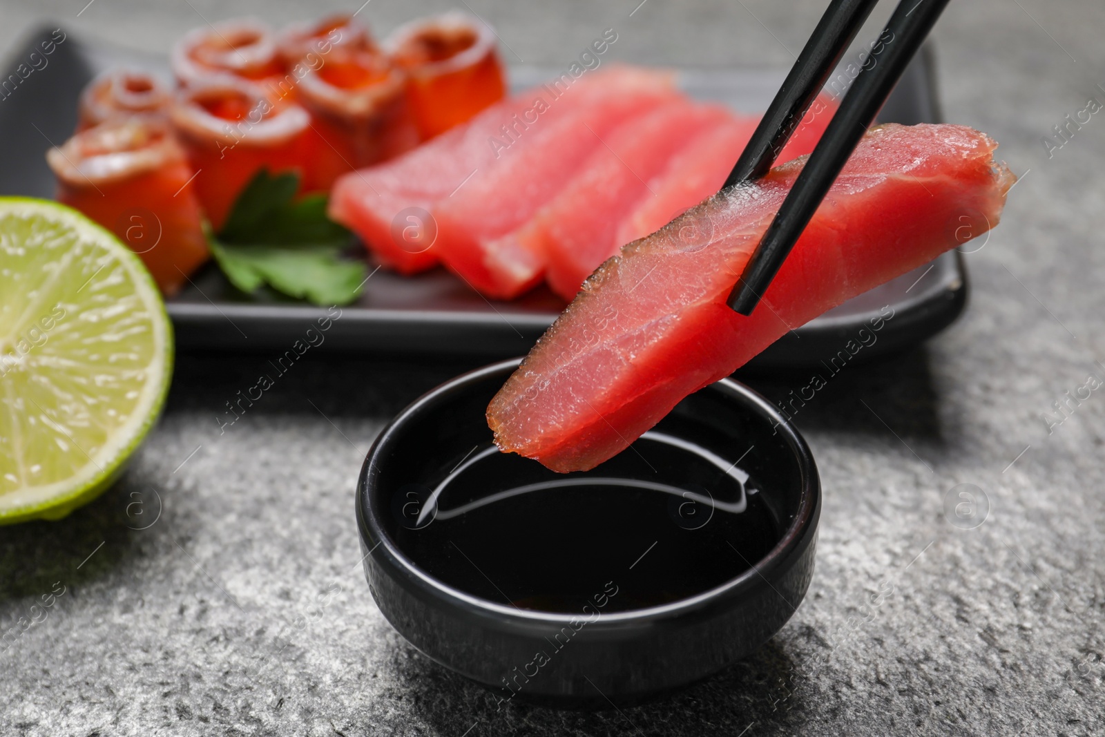 Photo of Chopsticks with raw tuna slice under soy sauce at grey table, closeup. Sashimi set served with lime