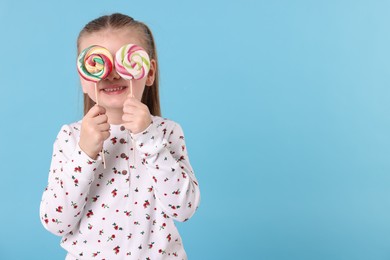 Photo of Happy little girl covering eyes with colorful lollipops on light blue background, space for text