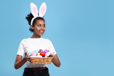 Photo of Happy African American woman in bunny ears headband holding wicker tray with Easter eggs on light blue background, space for text