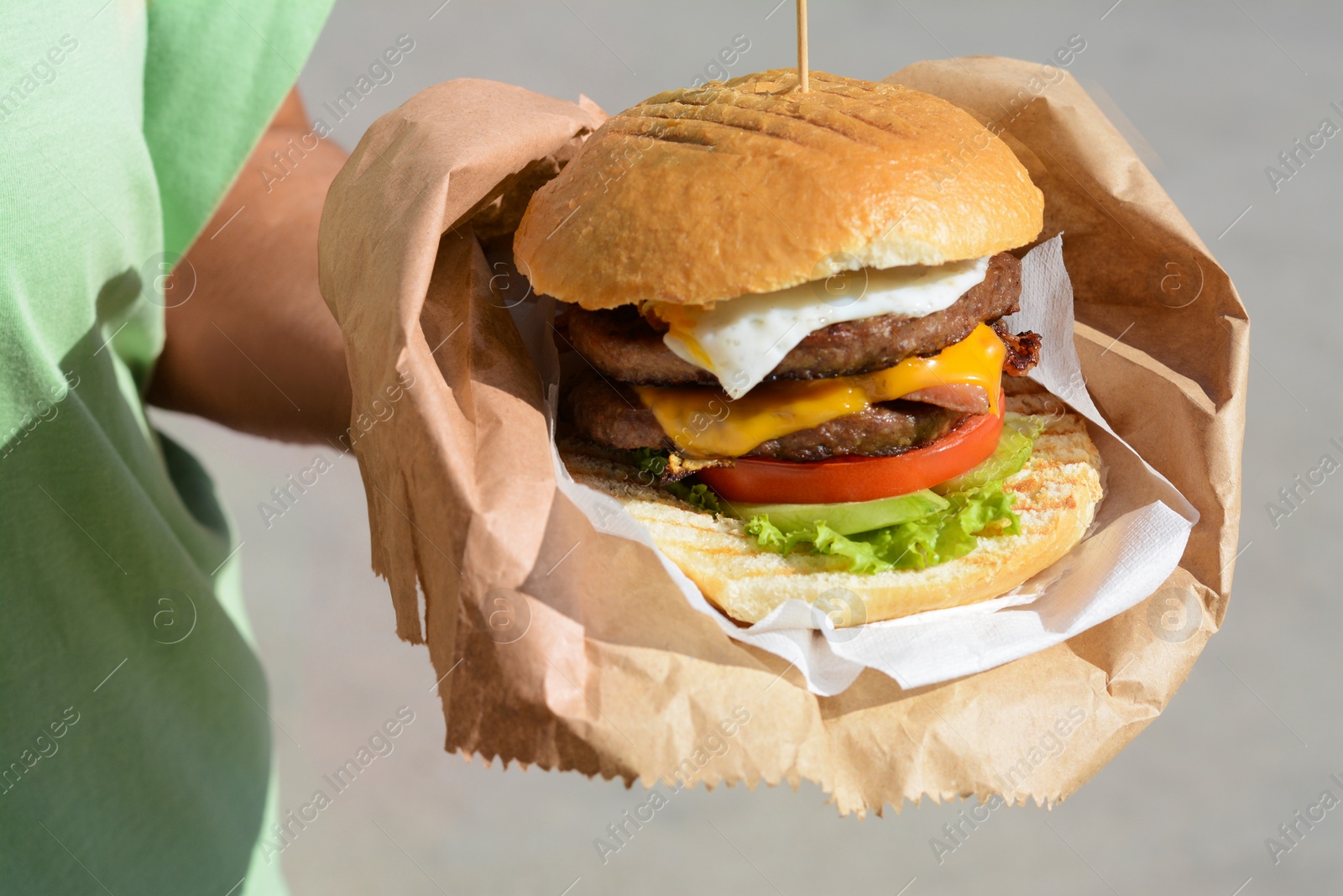 Photo of Woman holding delicious burger in paper wrap outdoors, closeup