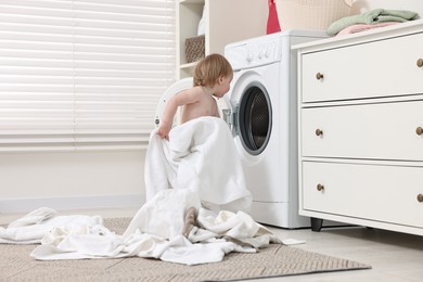 Photo of Little girl pulling baby clothes out of washing machine indoors