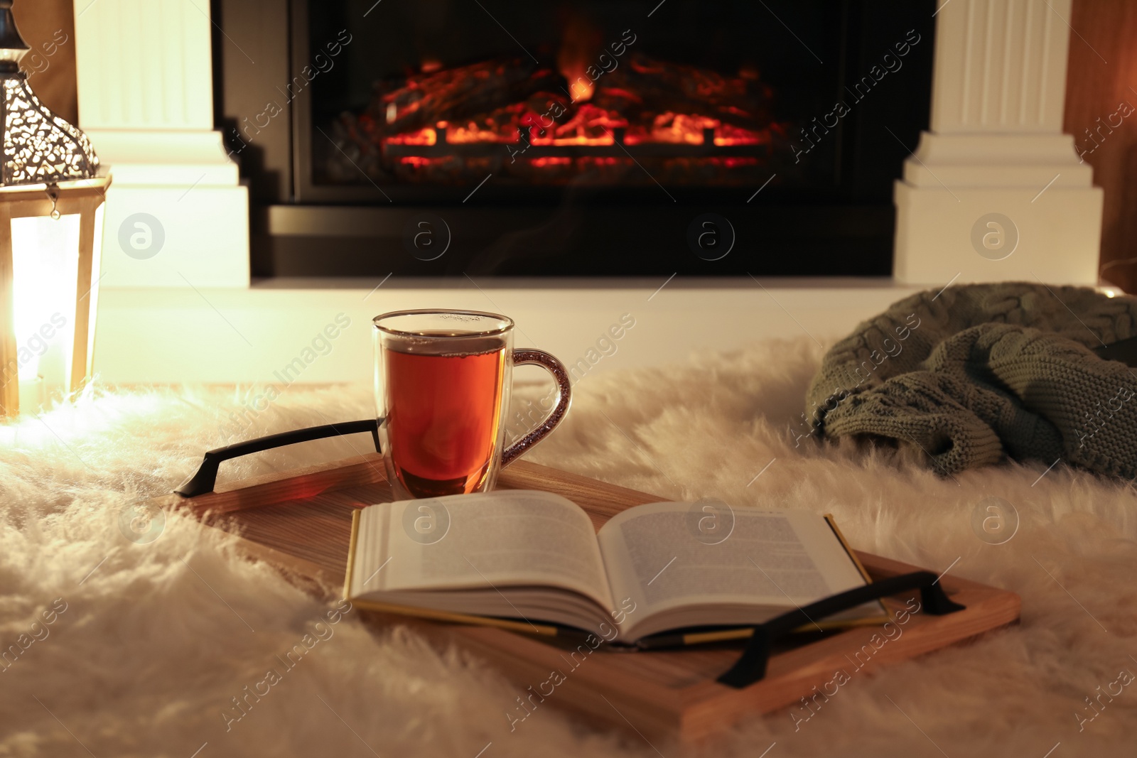 Photo of Cup of tea and book on fuzzy rug near fireplace at home. Cozy atmosphere