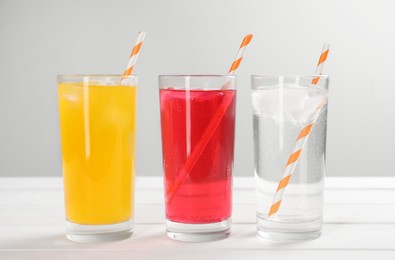 Glasses of different refreshing soda water with ice cubes and straws on white wooden table against light grey background