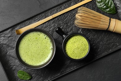 Cup of fresh matcha tea, bamboo whisk, spoon and green powder on black table, top view