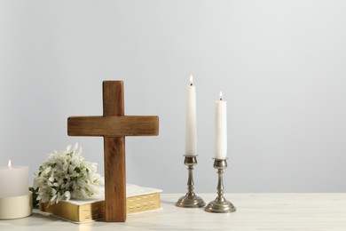 Burning church candles, wooden cross, Bible and flowers on white table. Space for text