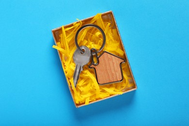 Key with trinket in shape of house and gift box on light blue background, top view. Housewarming party