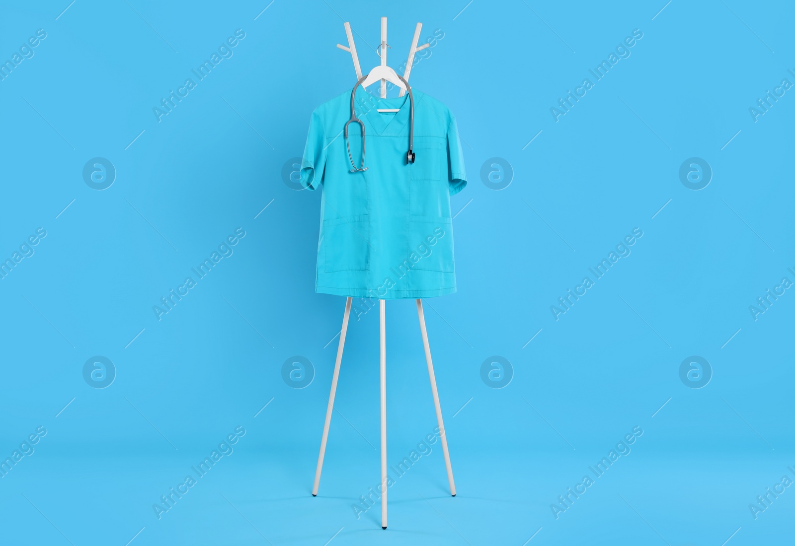 Photo of Turquoise medical uniform and stethoscope hanging on rack against light blue background