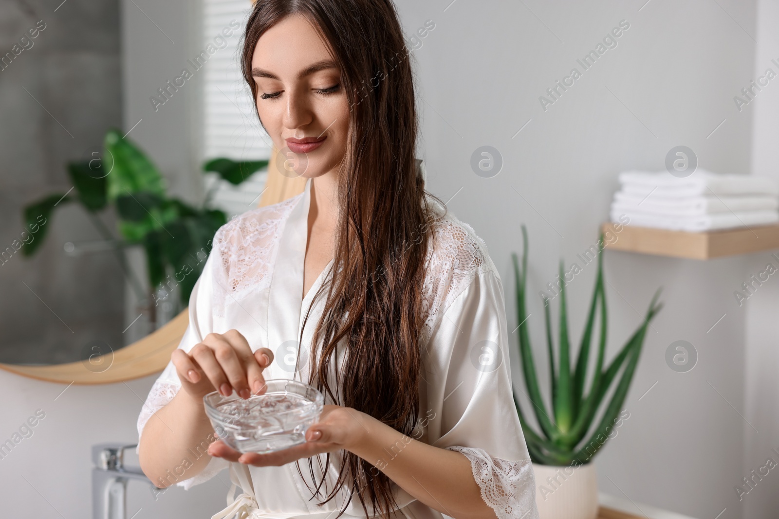 Photo of Young woman holding bowl of aloe hair mask in bathroom