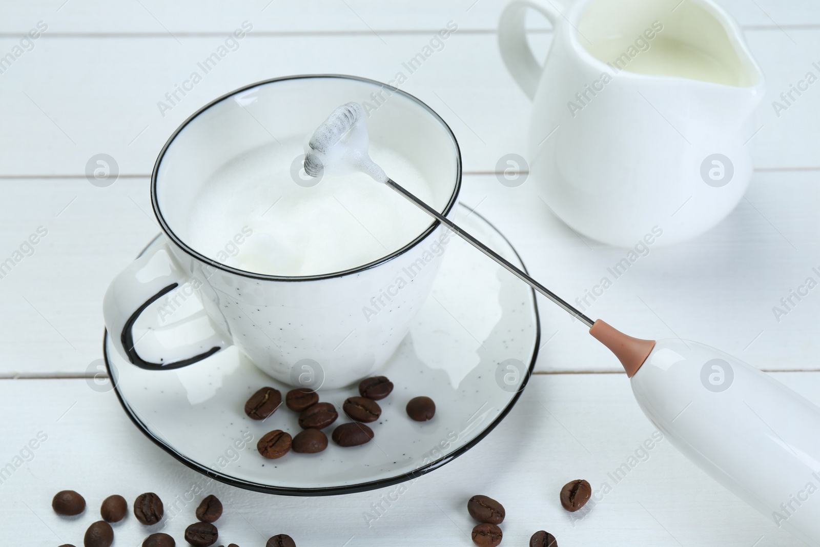 Photo of Mini mixer (milk frother), whipped milk and coffee beans on white wooden table, closeup