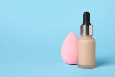 Bottle of skin foundation and sponge on light blue background, space for text. Makeup product