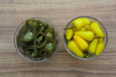 Glass bowls of pickled green and yellow jalapeno peppers on wooden table, flat lay