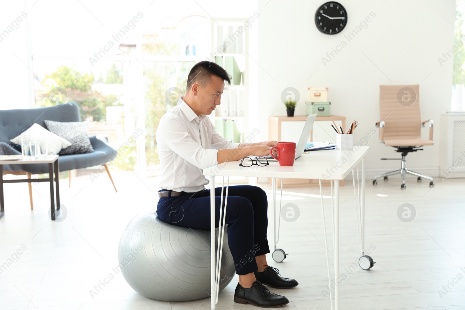 Photo of Young businessman sitting on fitness ball and using laptop in office. Workplace exercises