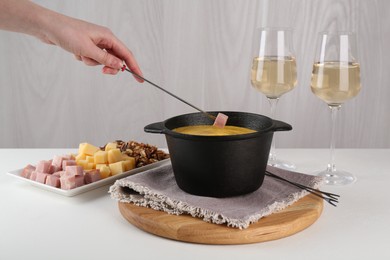 Photo of Dipping piece of ham into fondue pot with tasty melted cheese at white table, closeup