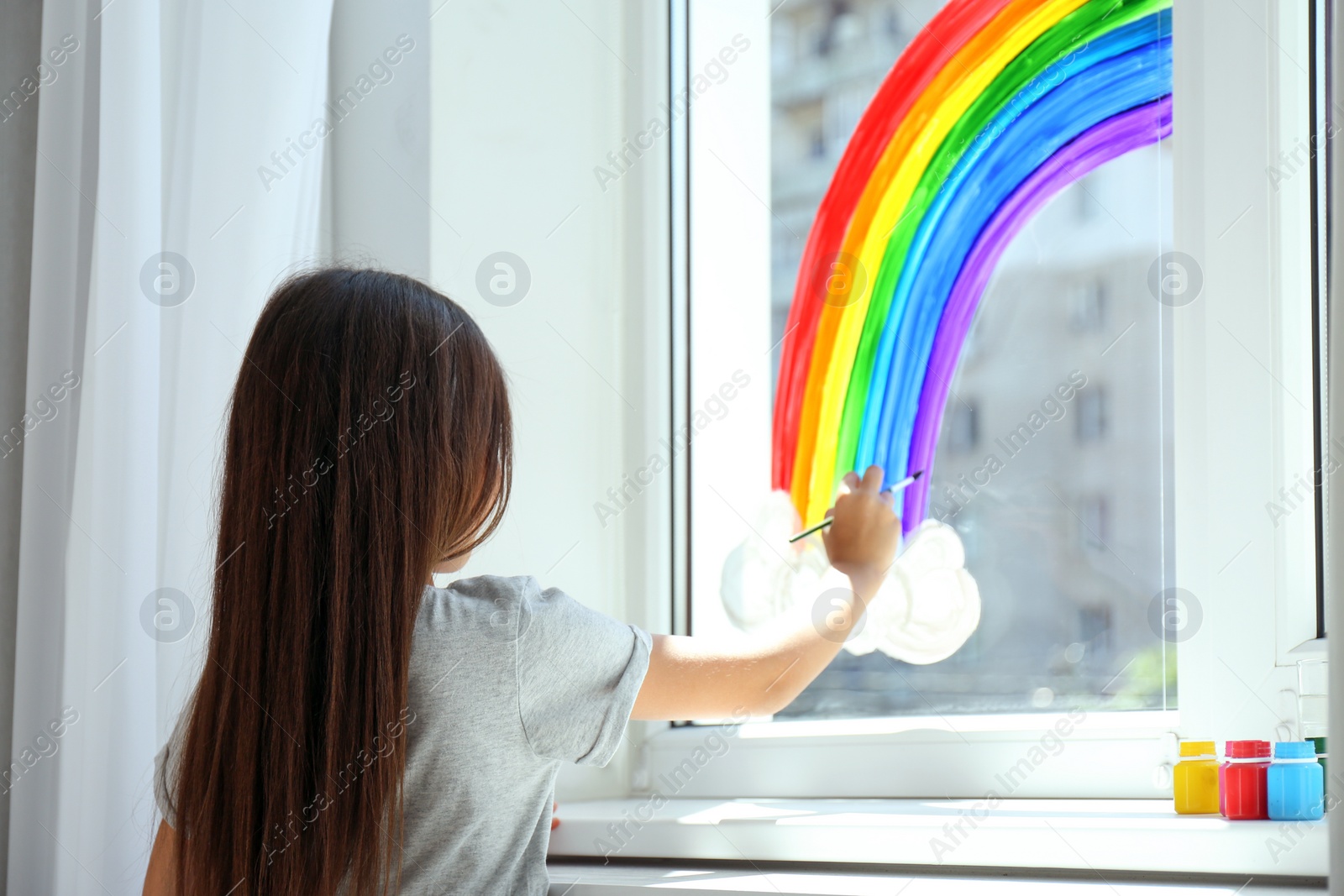 Photo of Little girl drawing rainbow on window indoors, back view. Stay at home concept
