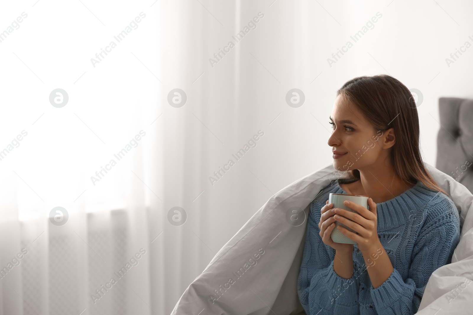 Photo of Young woman in warm sweater with hot drink on bed at home. Space for text