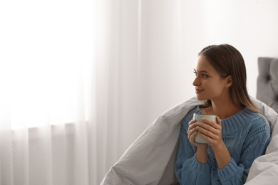 Photo of Young woman in warm sweater with hot drink on bed at home. Space for text