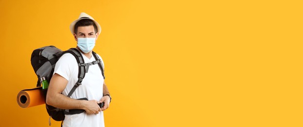 Male tourist in protective mask with travel backpack on yellow background, space for text