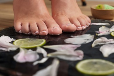 Photo of Woman soaking her feet in plate with water, flower petals and lime slices, closeup. Pedicure procedure