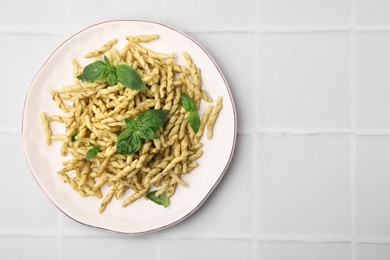 Plate of delicious trofie pasta with pesto sauce and basil leaves on white tiled table, top view. Space for text