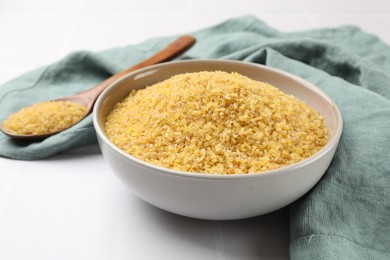 Bowl and spoon with raw bulgur on white tiled table