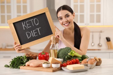 Photo of Happy woman holding chalkboard with words Keto Diet near different products in kitchen