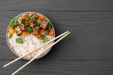 Bowl of rice with fried tofu, broccoli and carrots on grey wooden table, top view. Space for text