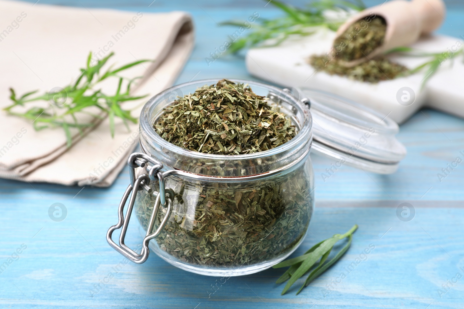 Photo of Jar of dry tarragon and green leaves on light blue wooden table, closeup
