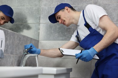 Photo of Smiling plumber with clipboard examining faucet in bathroom