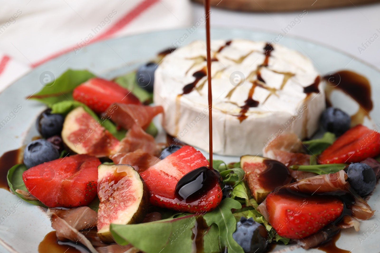 Photo of Pouring balsamic vinegar onto fresh salad with brie cheese, figs and berries on plate, closeup