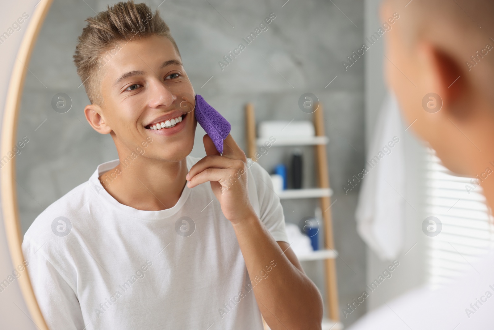 Photo of Happy young man washing his face with sponge near mirror in bathroom