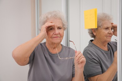 Photo of Forgetful senior woman near mirror with reminder note indoors. Age-related memory impairment