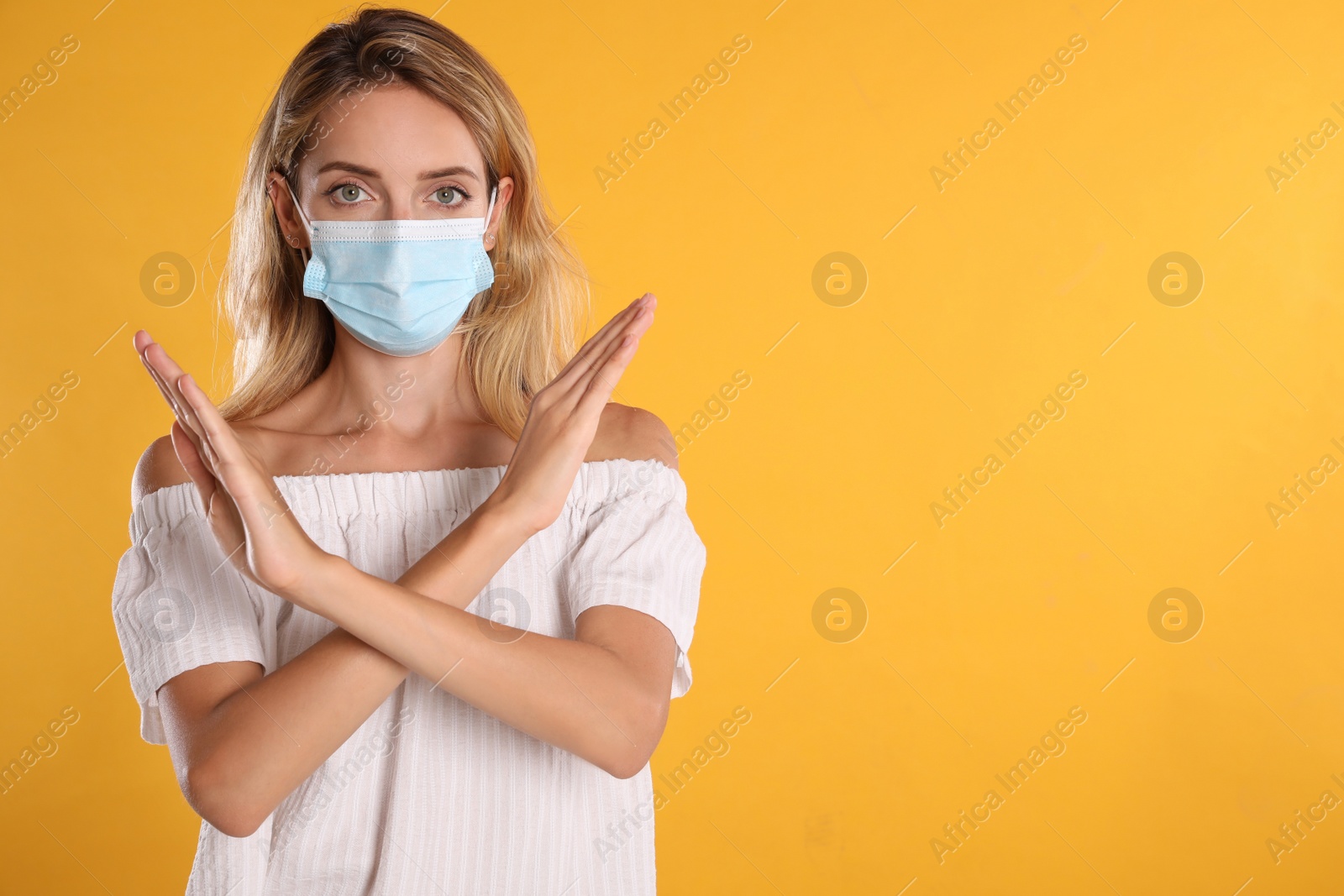Photo of Woman in protective face mask showing stop gesture on yellow background, space for text. Prevent spreading of coronavirus