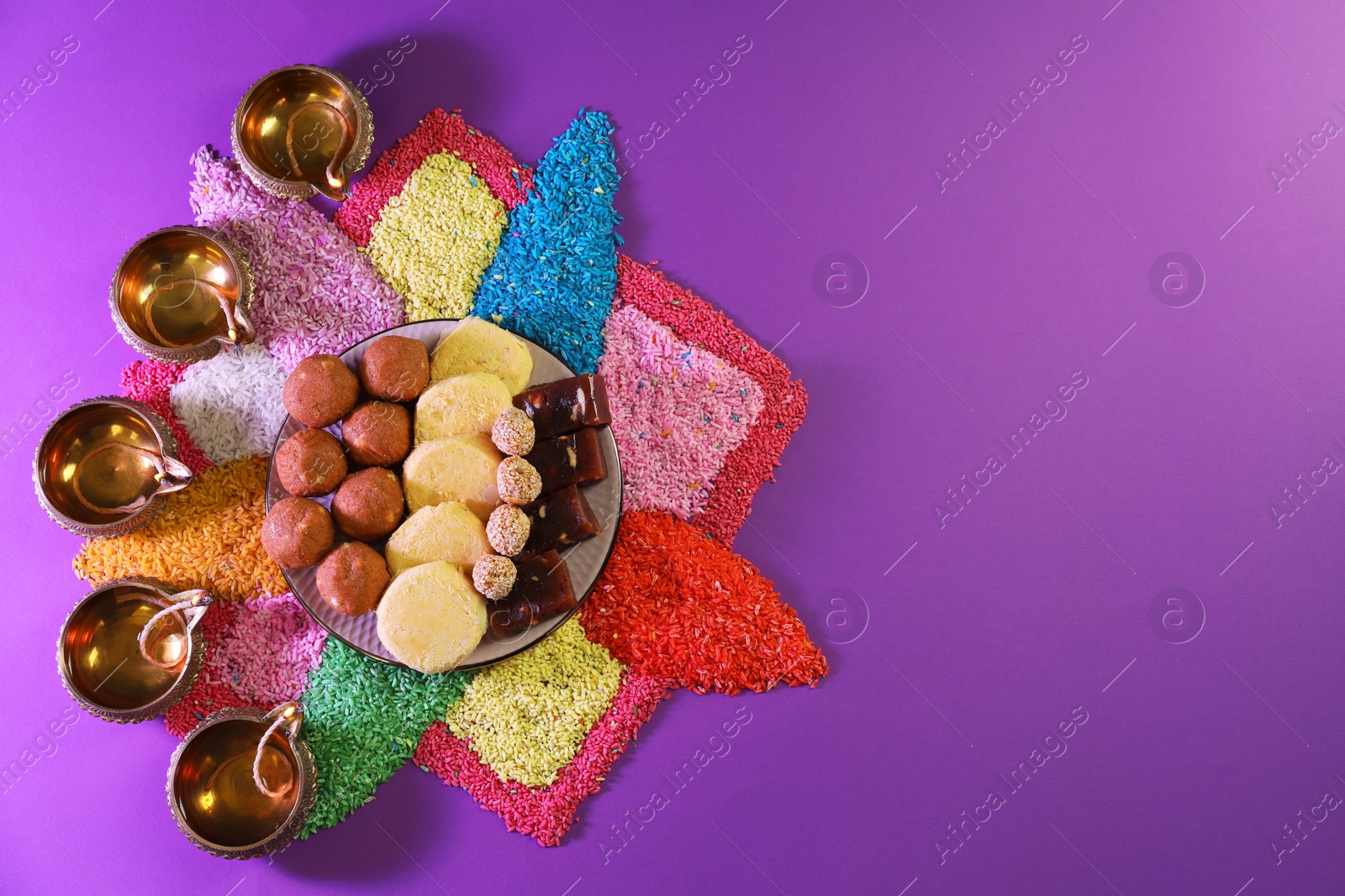 Photo of Diwali celebration. Tasty Indian sweets, colorful rangoli and diya lamps on violet background, flat lay. Space for text