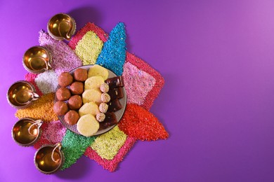 Photo of Diwali celebration. Tasty Indian sweets, colorful rangoli and diya lamps on violet background, flat lay. Space for text