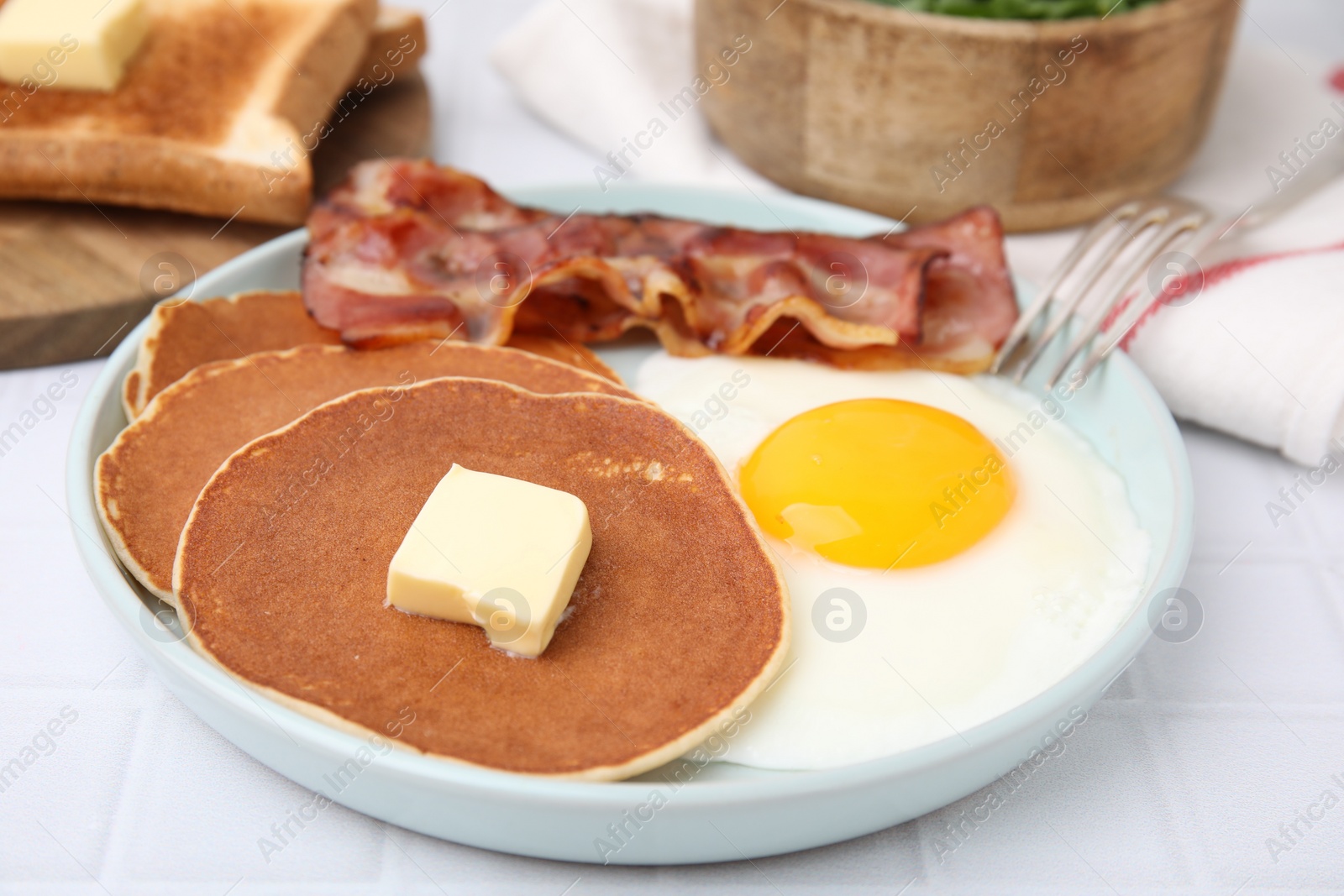 Photo of Tasty pancakes served with fried egg and bacon on white tiled table, closeup