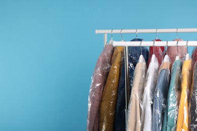 Photo of Dry-cleaning service. Many different clothes in plastic bags hanging on rack against light blue background, space for text