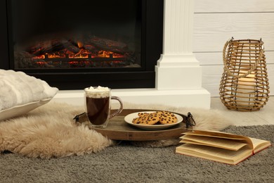 Open book, hot drink and cookies near decorative fireplace in room. Cozy home atmosphere