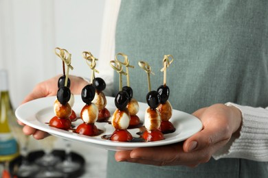 Photo of Woman holding plate of tasty canapes with black olives, mozzarella and cherry tomatoes indoors, closeup