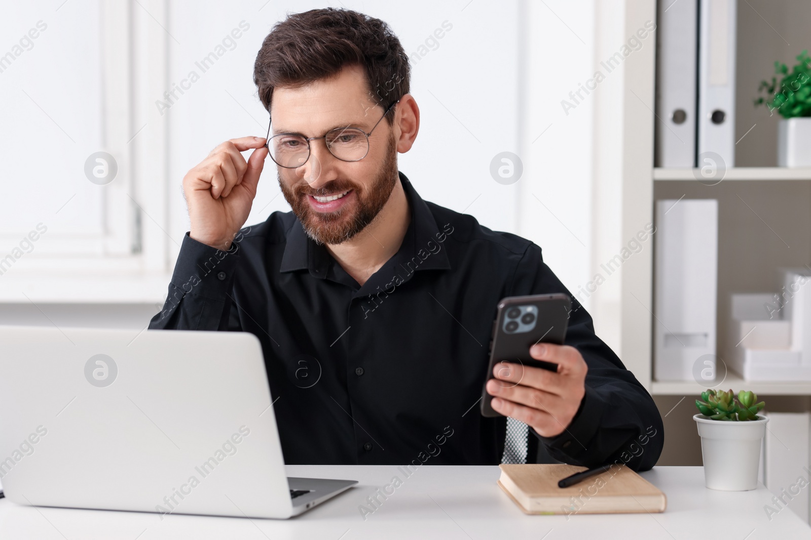 Photo of Smiling man with smartphone using laptop at table in office