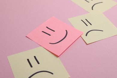 Choice concept. Sticky note with happy emoticon among beige papers with sad emojis on pink background, closeup