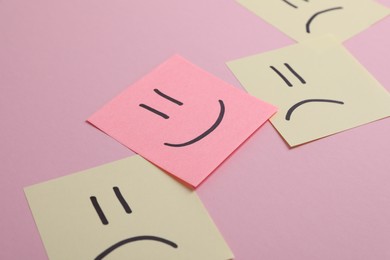 Photo of Choice concept. Sticky note with happy emoticon among beige papers with sad emojis on pink background, closeup