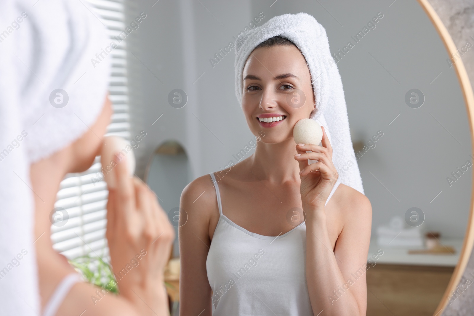 Photo of Happy young woman washing her face with sponge near mirror in bathroom