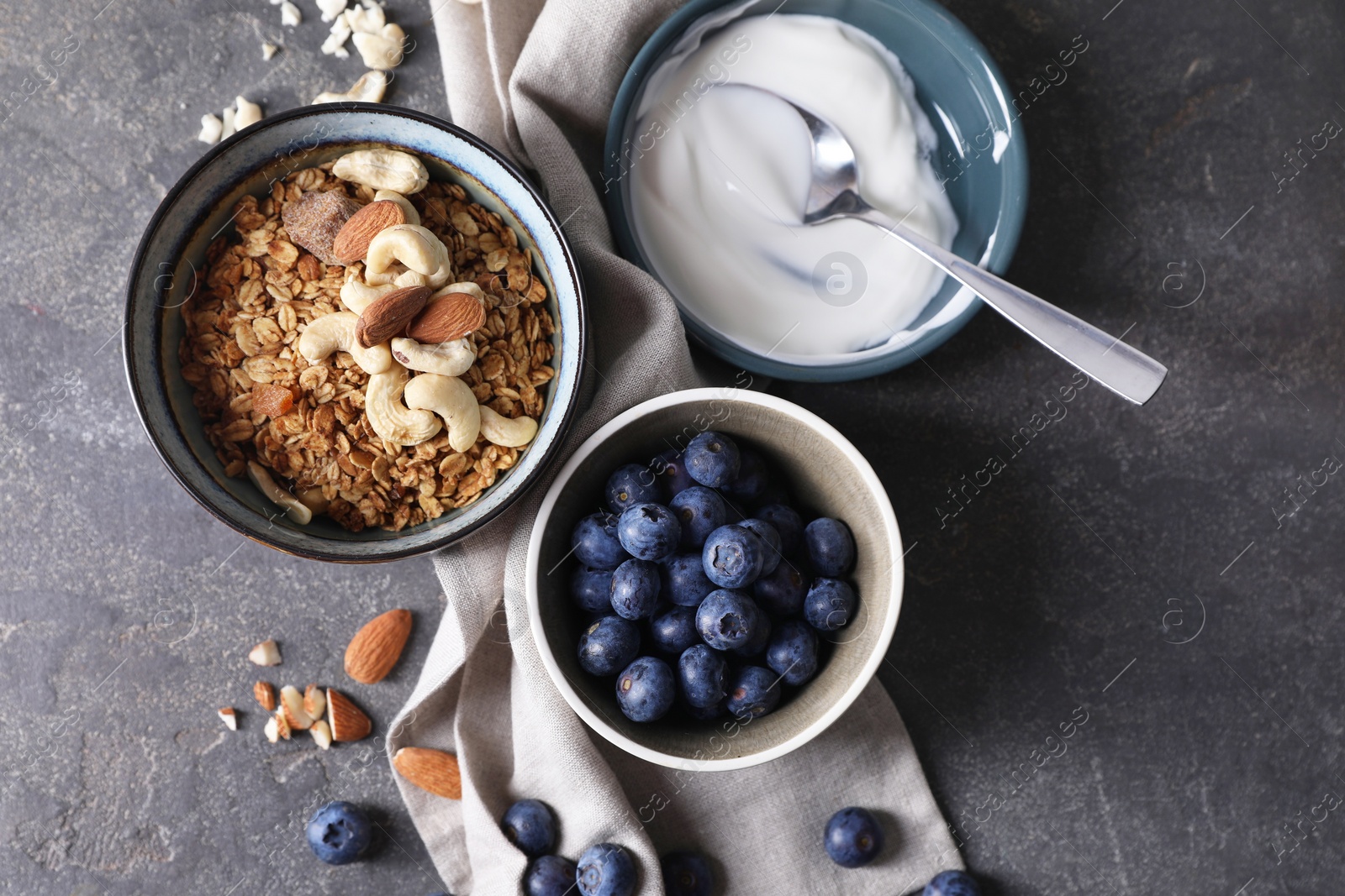 Photo of Tasty granola in bowl, blueberries, yogurt and spoon on gray textured table, flat lay